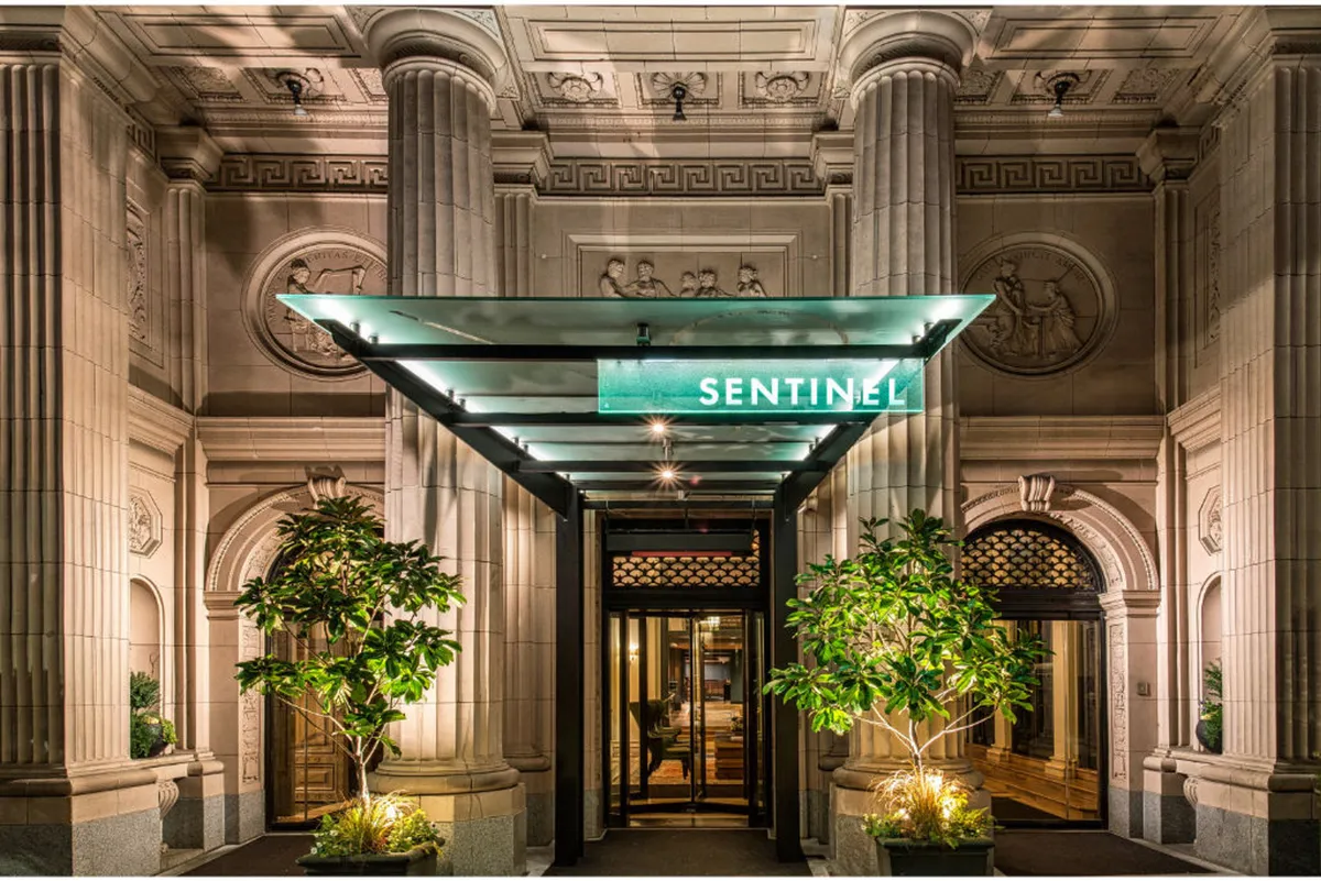 The exterior entrance of Hotel Sentinel, marked by an elegant awning with the 'SENTINEL' sign, ornate architectural details, and lush planters, offers a sophisticated urban welcome. The greenery hints at easy access to outdoor spaces for pet walks.