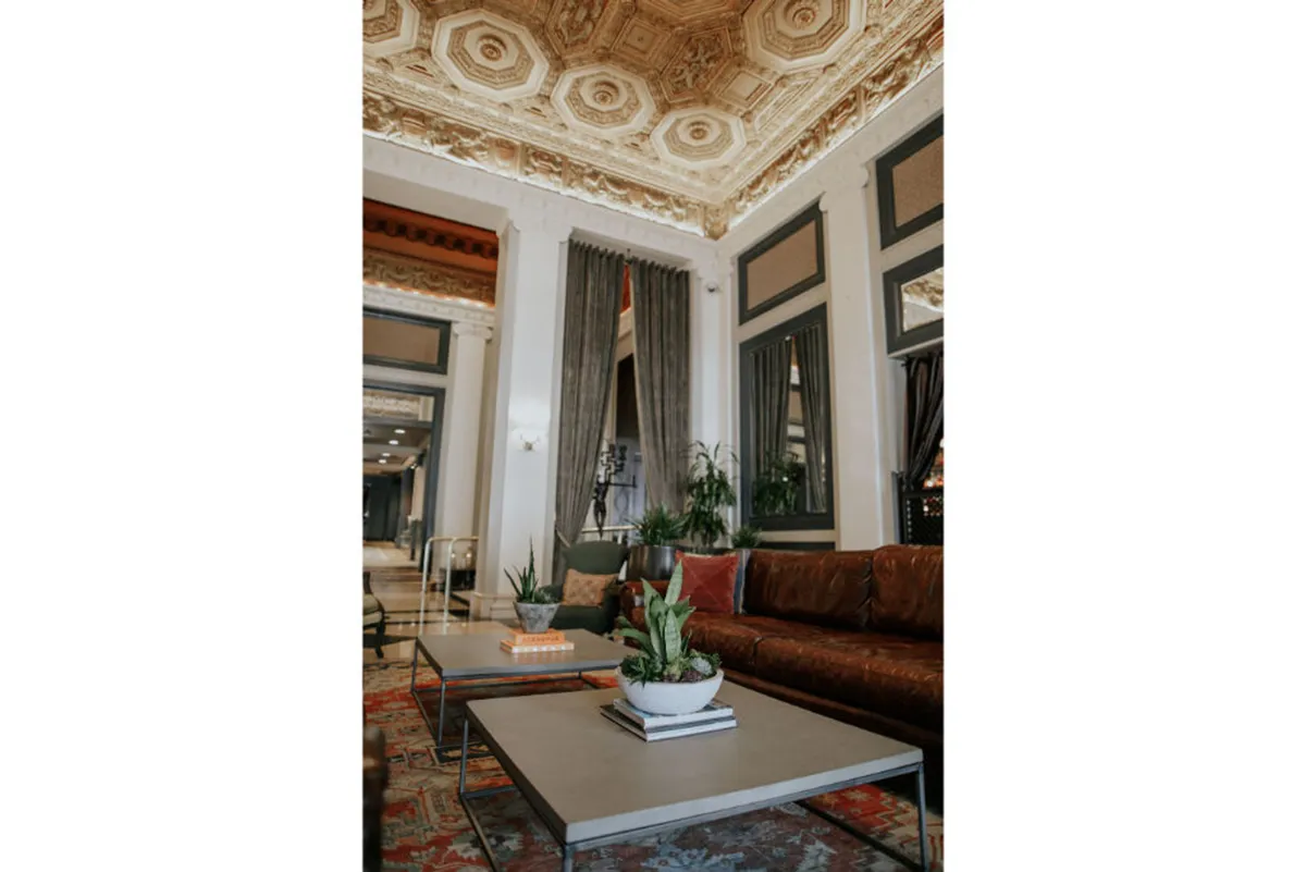 This image features an ornate ceiling above a luxurious sofa, complemented by coffee tables adorned with plants, all set within a sophisticated and comfortable interior. The arrangement invites relaxation, reading, or casual conversation, echoing the ambiance of a thoughtfully designed guest room.