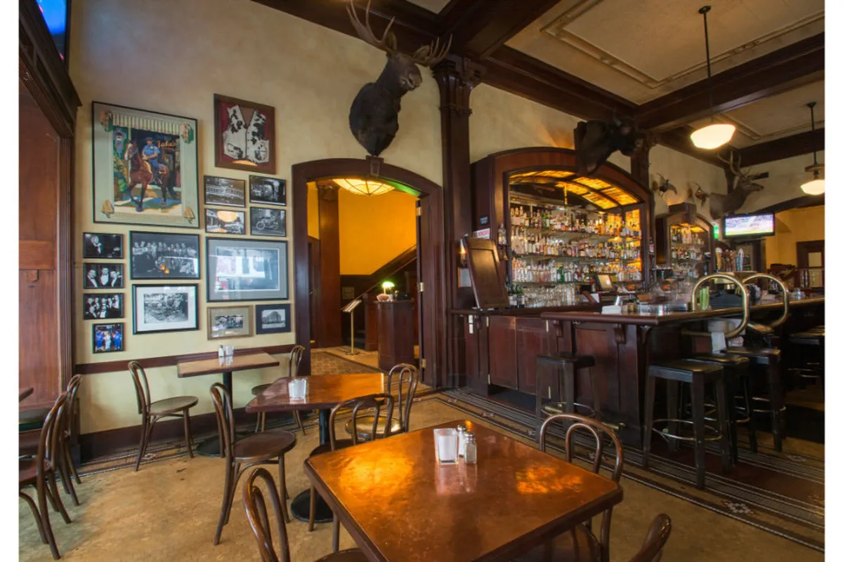 The scene features a vintage-inspired bar counter adorned with liquor bottles, vintage photographs, and decorative moose heads, surrounded by wooden tables and chairs. Guests are seen socializing, enjoying drinks, dining, and watching sports, evoking a warm, nostalgic mood.