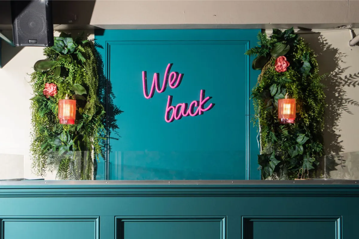 A lively interior scene featuring neon signage, abundant greenery, and decorative lighting. The setting is vibrant and social, with groups of people relaxing and celebrating together. The playful, welcoming mood creates an inviting atmosphere for gatherings and casual fun.