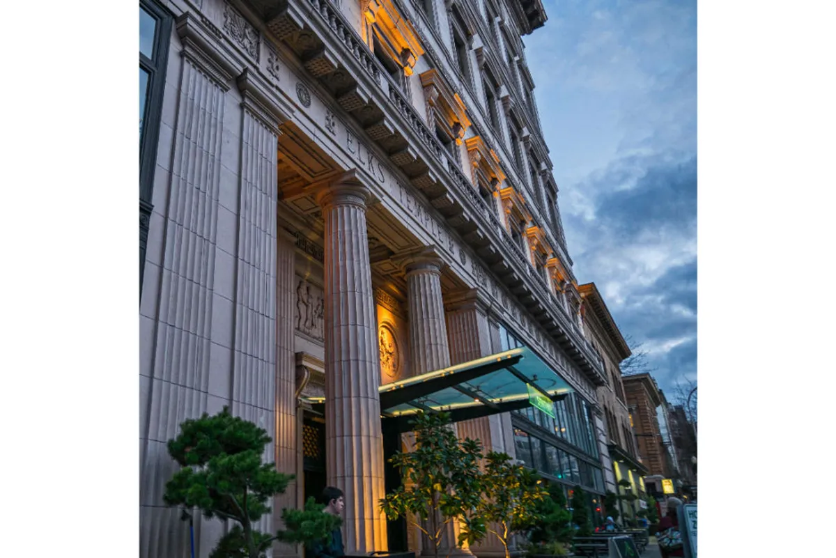 An exterior view highlighting the grand facade of a classic building with stately columns, illuminated signage, and lush greenery. Guests are seen dining outdoors and socializing at the entrance, evoking a sense of community and urban vibrancy.