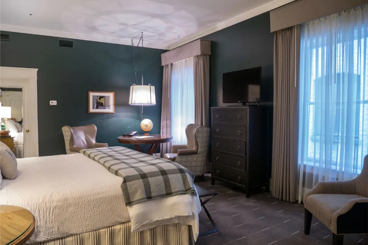 The image showcases a beautifully appointed hotel room featuring a plush bed with a stylish patterned throw blanket, a warm-lit bedside lamp atop a wooden table, and an inviting sitting area with two elegant armchairs. The overall ambiance is serene and comfortable, perfect for relaxation or intimate conversation.