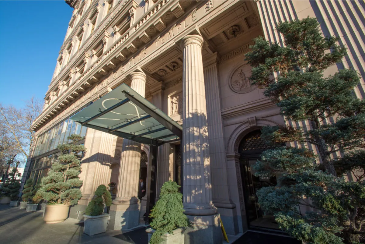 This image showcases the sophisticated entrance of Hotel Sentinel, featuring ornate columns, a glass canopy, manicured trees, and an inviting doorway. The scene suggests a sense of arrival and anticipation, perfectly capturing the transition from urban adventure to luxurious respite.